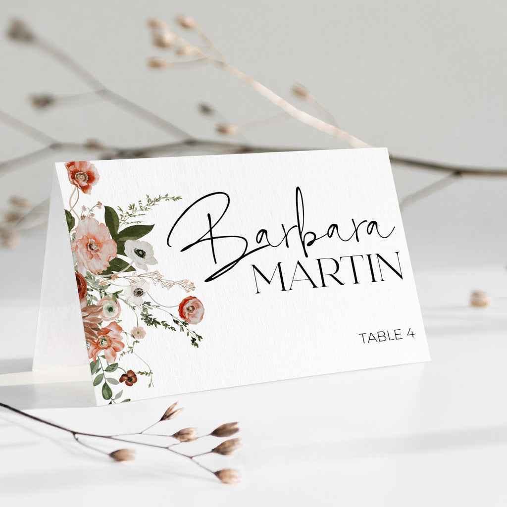 A tent folded place card with a floral design and the name written on it, displayed on a white background with dried flowers around.