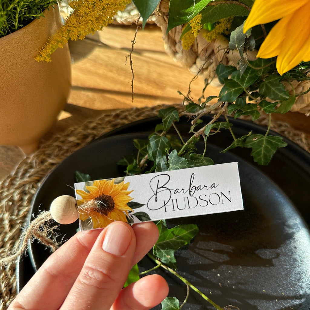 A tent folded place card with a sunflower design, personalized with a name, displayed on a table with a floral arrangement in the background.