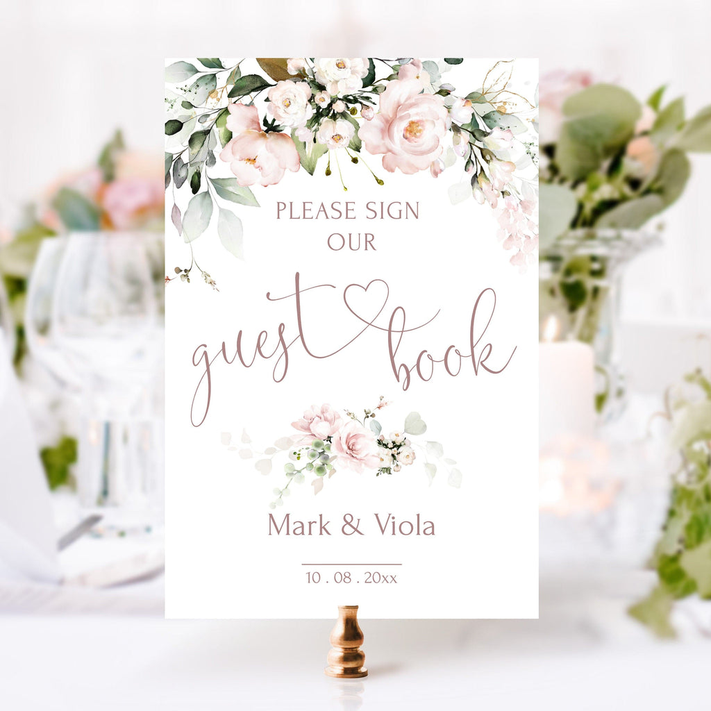 A blush pink sign with floral design and the text 'Please sign our guest book' printed on it, placed on a table with decorations in the background.