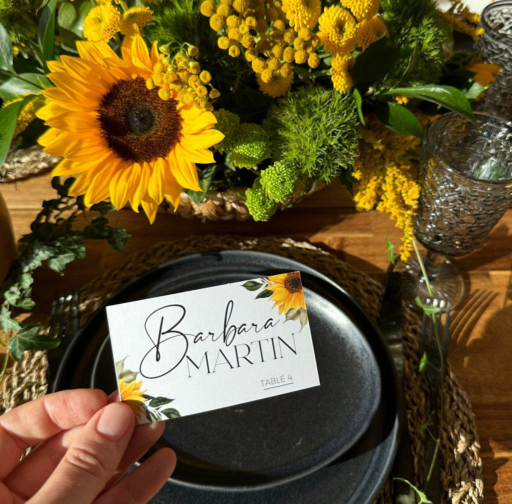 A hand holding a folded place card with the name 'Barbra Martin' written on it, with a sunflower design in the background.