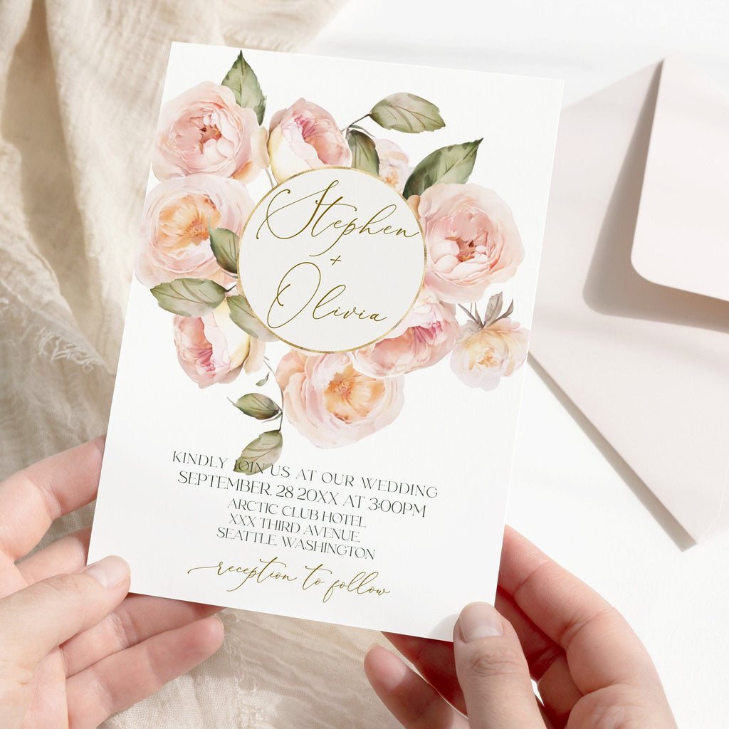 A hand holding a white wedding invitation card with a floral design featuring peach fuzz roses and the names 'Stephanie' and 'Oliver' on the front. The back of the card is visible with a text area for personalization.