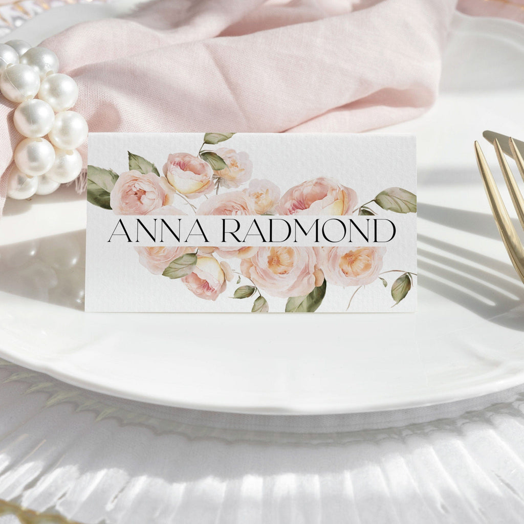 A tent folded place card with a floral design featuring peach fuzz roses and the name 'Anna Radmond' printed on it, placed on a white plate with a pink napkin and decorative flowers in the background.