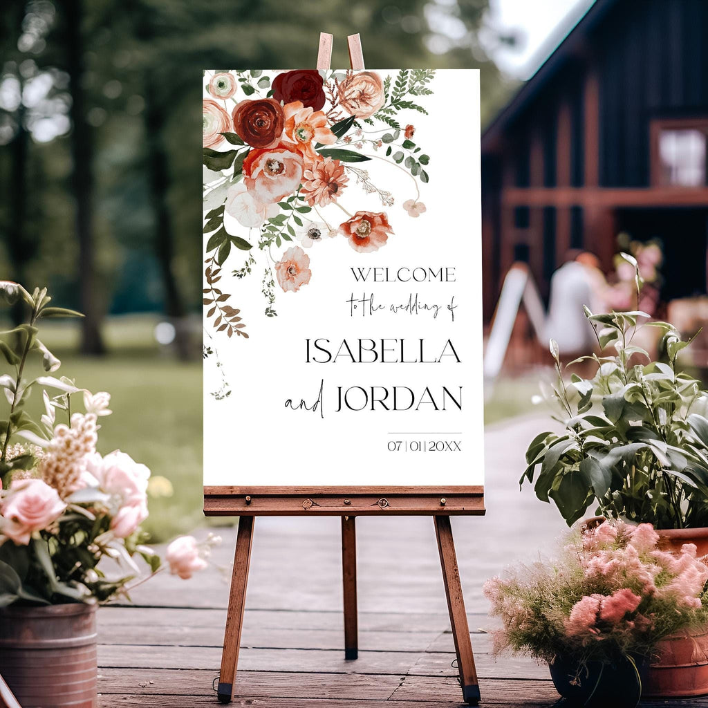 A welcome sign for a wedding with a floral design, the sign is displayed on an easel outdoors, surrounded by floral arrangements.