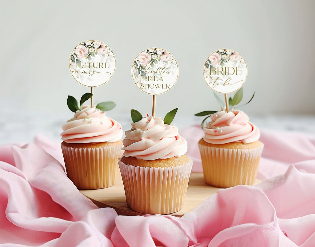 Three cupcakes with blush pink frosting, each topped with a floral cupcake topper with a different text design, presented on a pink fabric surface.